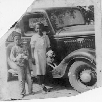 Two children stand in front of a car, each holding a dog.